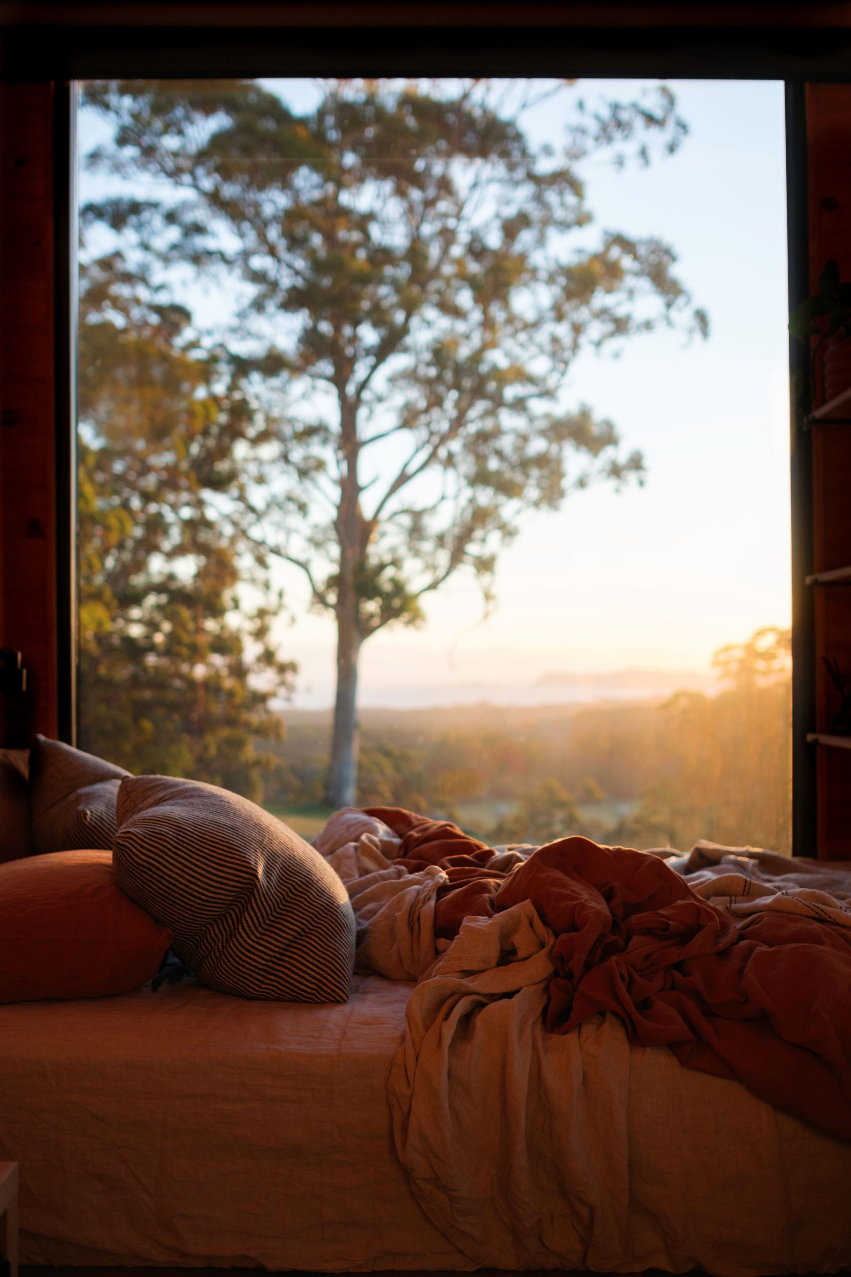 Unmade bed in one-bedroom ocean view cabin in Denmark WA with window view of early sunset over landscape