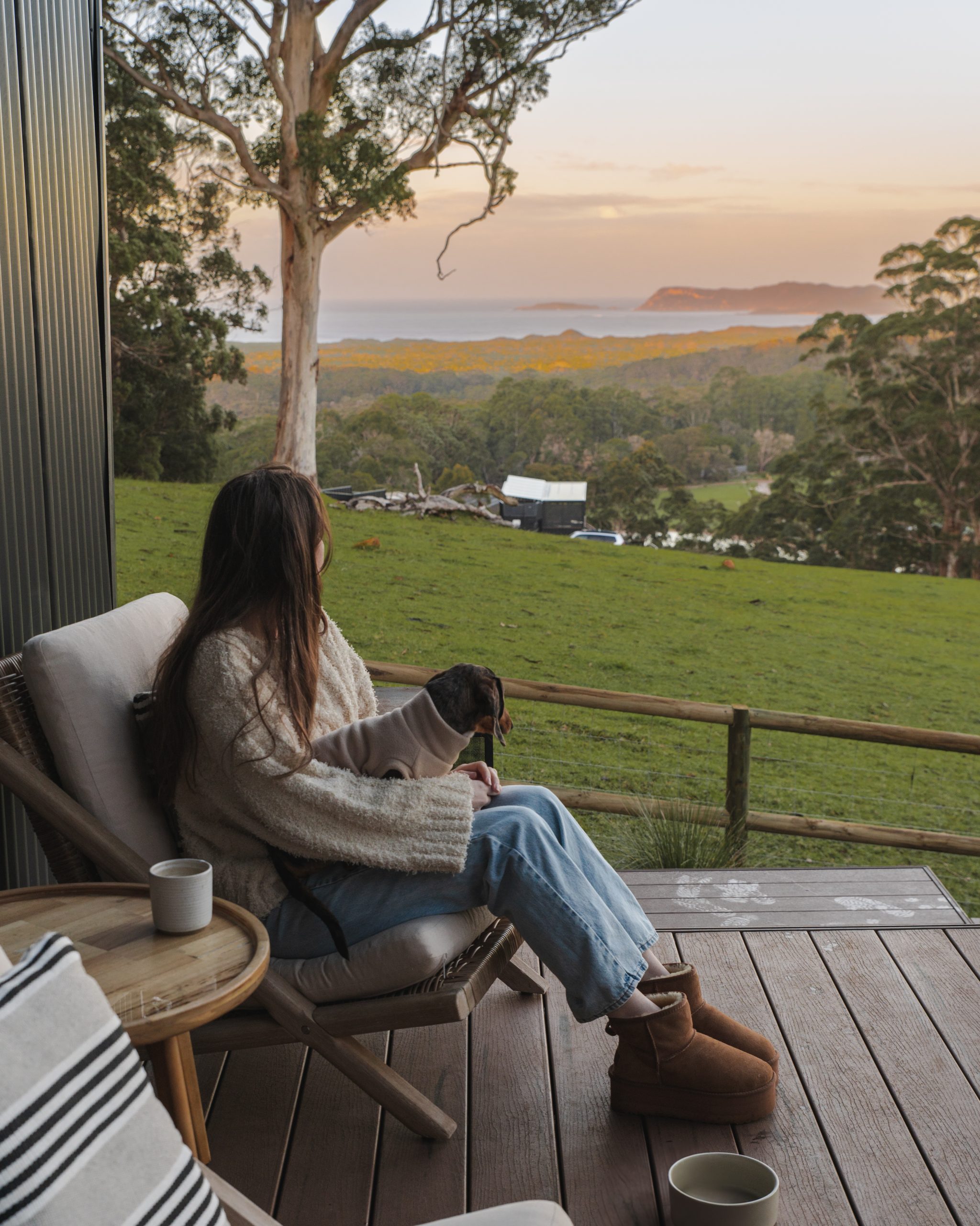 Woman on balcony of ocean view one-bedroom cabin in Denmark WA with dog on lap overlooking coastal landscape